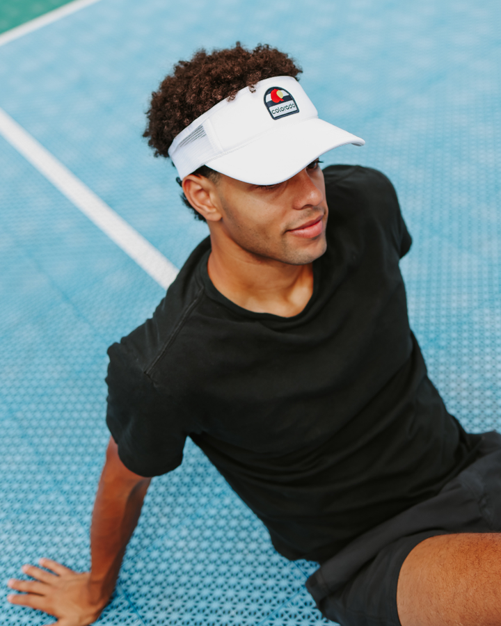 A man wearing a white visor and a black t-shirt sits on a blue tennis court. The trucker visor features the red and blue colorado logo on the front.
